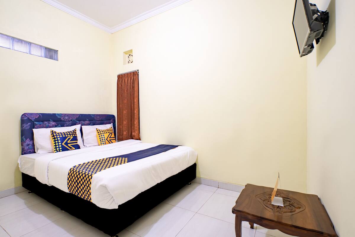 A neatly made bed with a patterned bedspread, a wooden nightstand with a lamp, and a window with blinds.