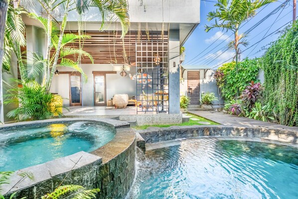 A modern outdoor pool area with a waterfall feature, surrounded by lush greenery and a contemporary building with a wooden door.