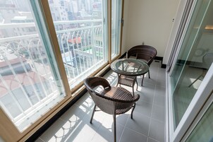 Un balcón con sillas de mimbre y una mesa de vidrio, con vistas a un paisaje urbano que incluye edificios y una carretera.