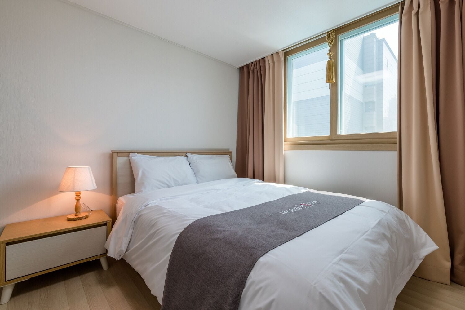 A neatly made bed with a grey blanket, a bedside lamp, and a window with curtains.