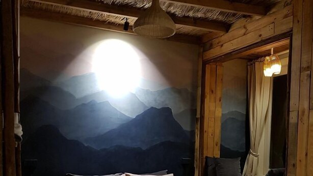 Een houten kamer met een bed, een muurschildering van bergen, een hanglamp en een raam met gordijnen.