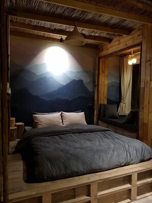 Una habitación de madera con una cama, un mural de montañas en la pared, una lámpara colgante y una ventana con cortinas.
