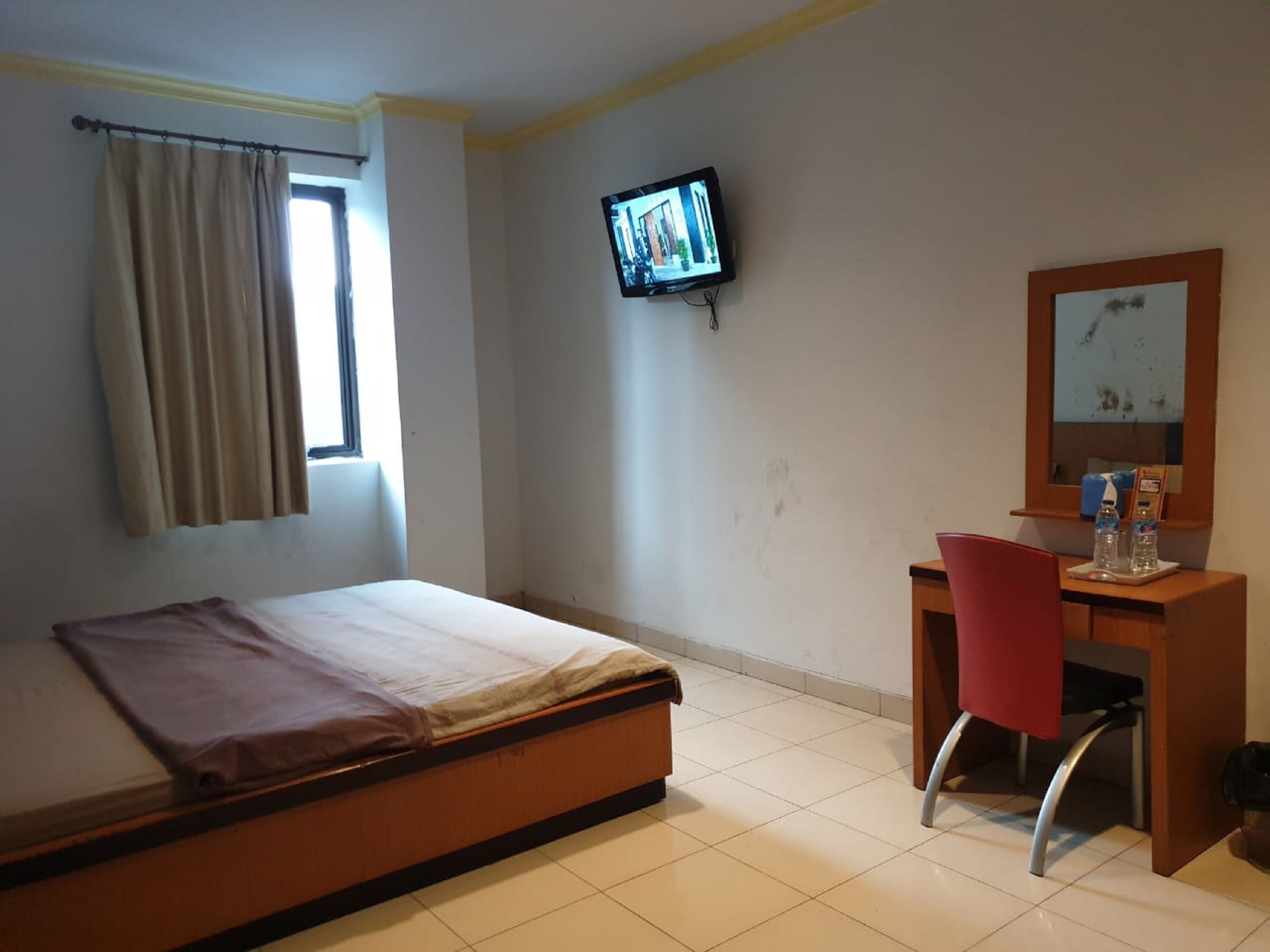 A hotel room with a bed, a desk with a chair, a television mounted on the wall, and a window with curtains.