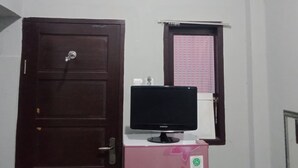 Ein Zimmer mit einer weißen Klimaanlage, einer braunen Tür, einem rosa Kühlschrank mit Aufkleber, einem Samsung-Monitor darauf und einem Fenster mit einem rosa gemusterten Rollo.