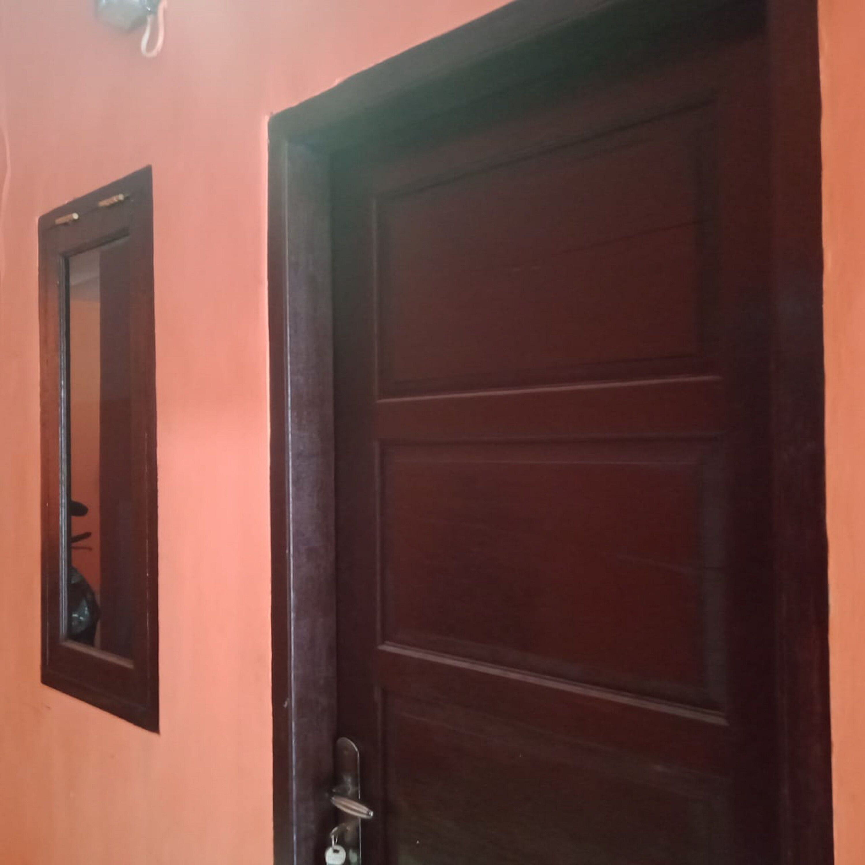Una puerta de madera con un ojo de cerradura y un pomo, un espejo pequeño en la pared y un interruptor de luz encima de la puerta.