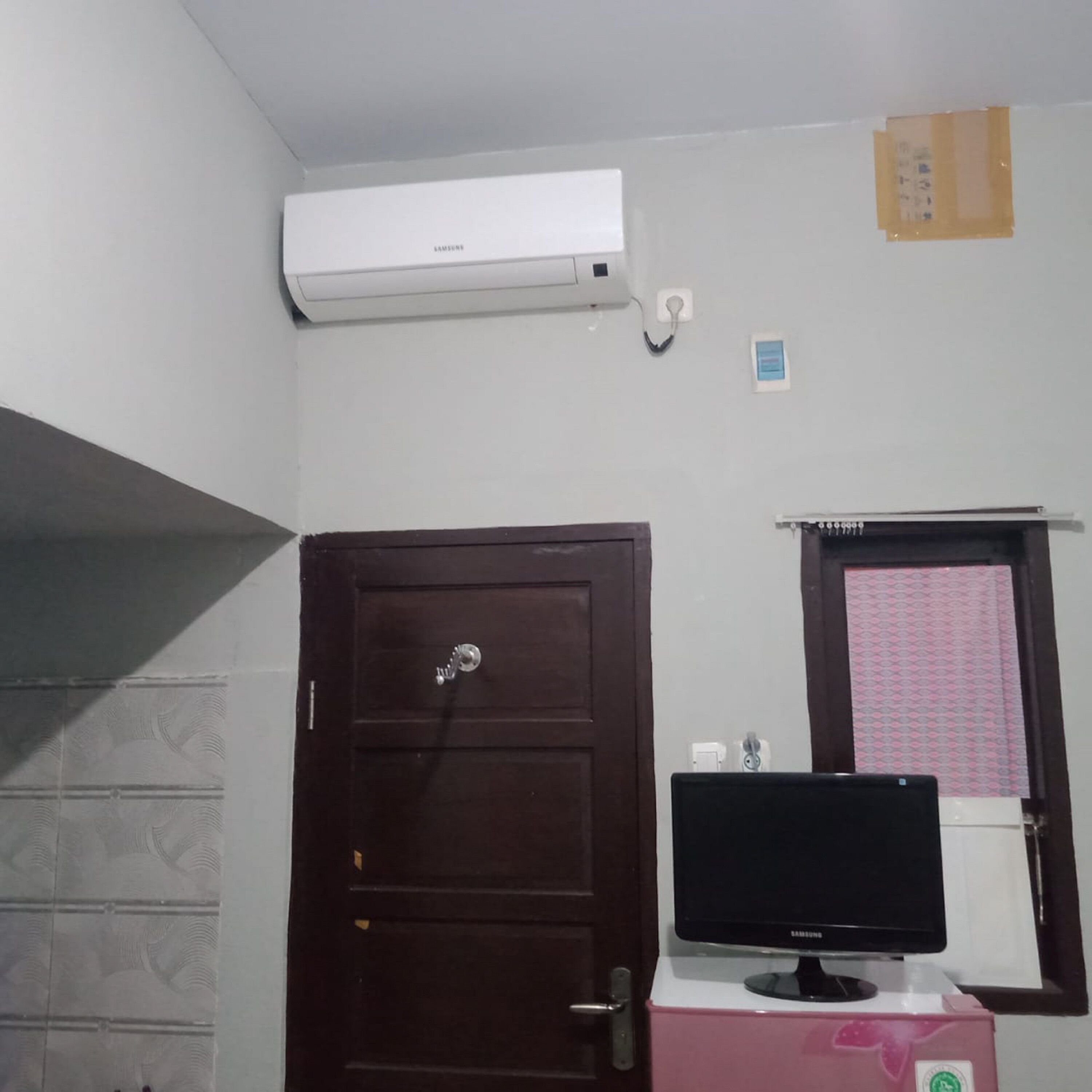 A room with a brown door, a wall-mounted air conditioner, a window with a pink patterned curtain, and a Samsung monitor on a pink stand.