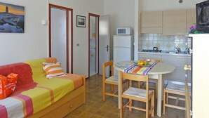 Een knus woongedeelte met een bank, een eettafel met stoelen, een keuken met fornuis en magnetron, en een deur die naar een andere kamer leidt.