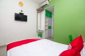 Habitación de hotel con una cama, un televisor de pantalla plana, una planta pequeña, un cubo de basura verde y una pared verde.