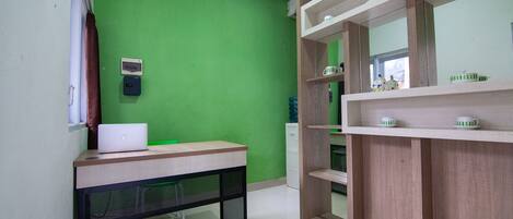 A room with a green wall, a wooden desk with a laptop, a white cabinet, and a shelf with cups.