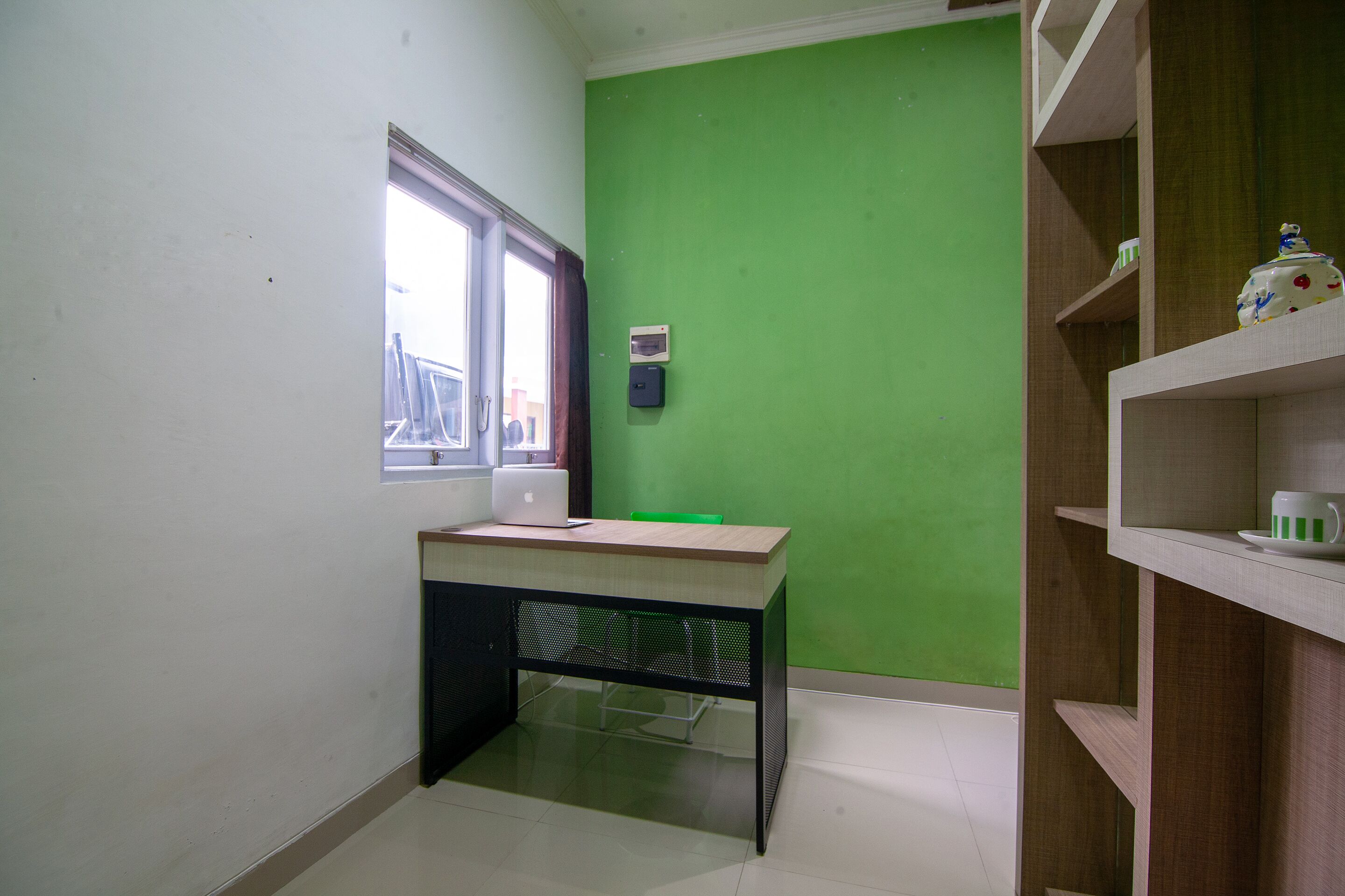 Una habitación con pared verde, ventana, escritorio con computadora portátil y estante con tetera.