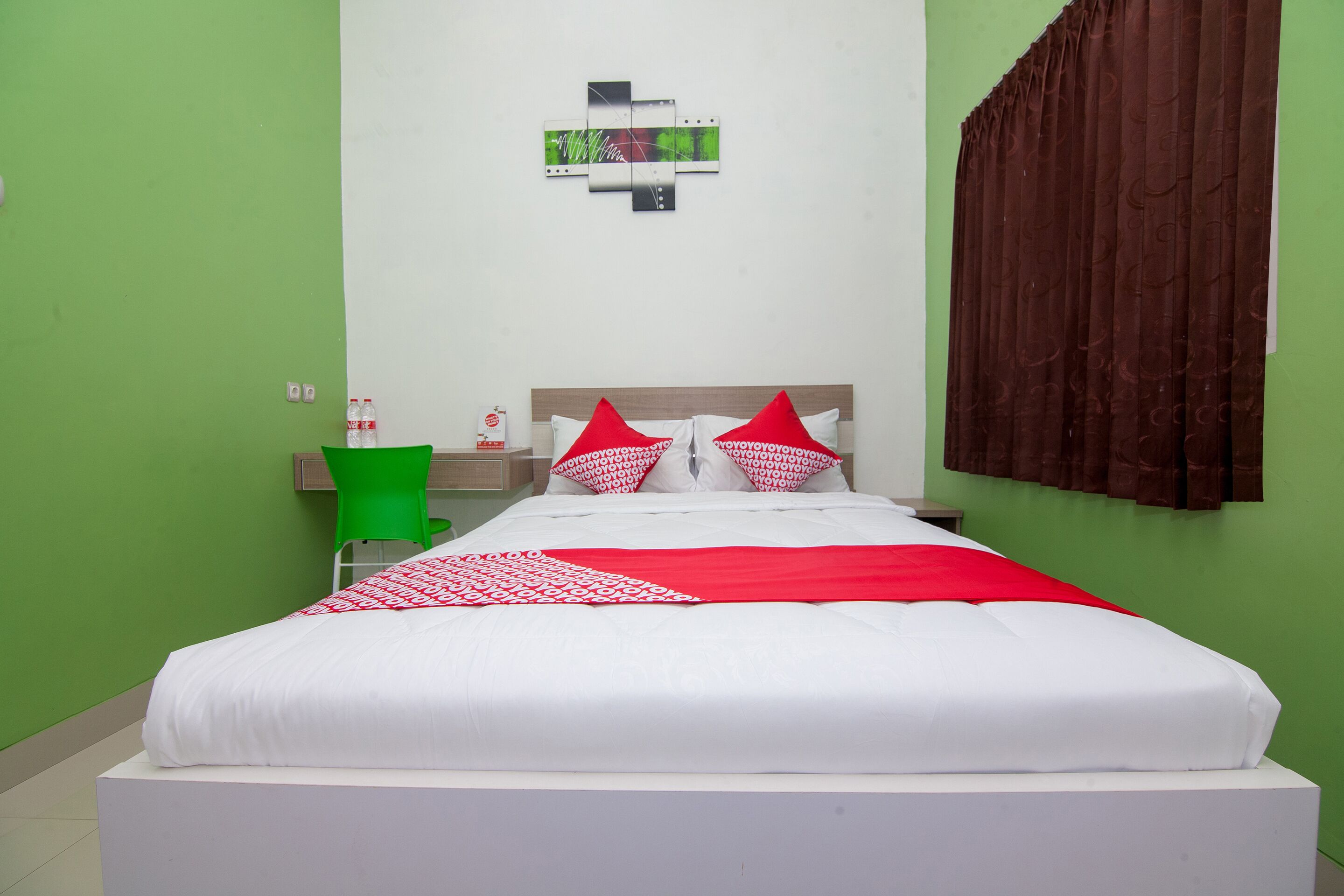 Una cama bien hecha con ropa de cama blanca y roja, una silla verde y una obra de arte colgada en la pared.