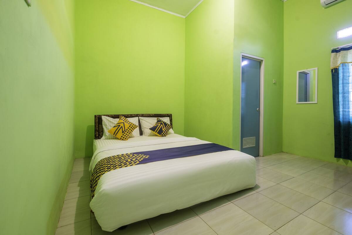 Een kamer met groene wanden, een bed met wit beddengoed en gekleurde kussens, een klein raam met spiegel en een deur.