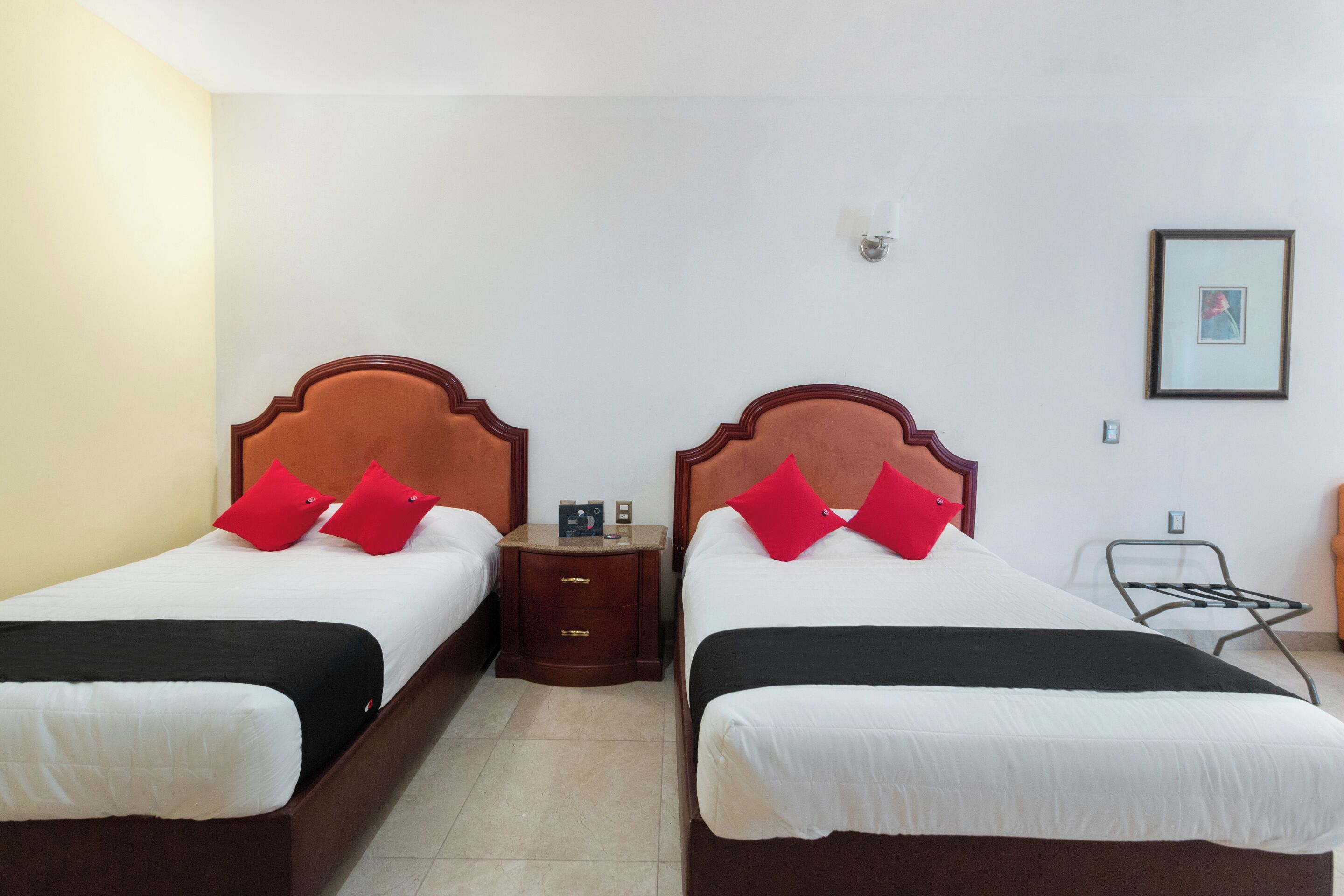 A hotel room with two beds, each with a wooden headboard, white bedding, and red pillows. There is a nightstand between the beds with a lamp and a small table.
