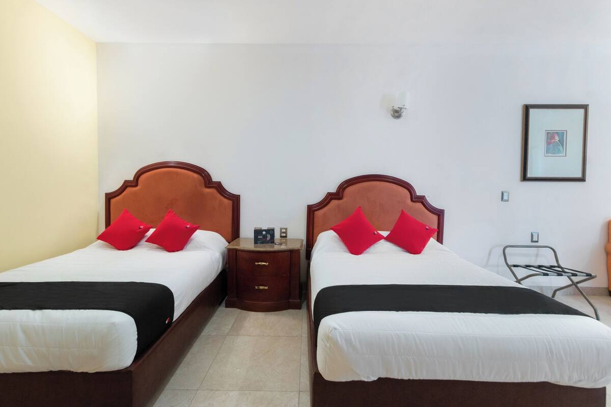 A hotel room with two beds, each with a wooden headboard, white bedding, and red pillows. There is a nightstand between the beds with a lamp and a small table.