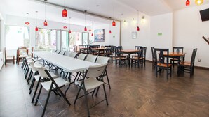 Uma sala de jantar ampla com mesas e cadeiras compridas, luminárias pendentes vermelhas e vista para o verde lá fora.