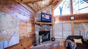 A cozy cabin interior with a stone fireplace, wooden walls, a mounted TV, and a large window with blinds.