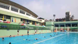 Une piscine dotée de plusieurs couloirs, d’une plateforme de plongeon et d’un bâtiment à la façade verdoyante, ornée de drapeaux.
