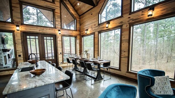 A spacious kitchen with a marble countertop, dining area with chairs, and large windows offering a view of the forest. - Across the Universe - Broken Bow/Hochatown- 3bd (Broken Bow)