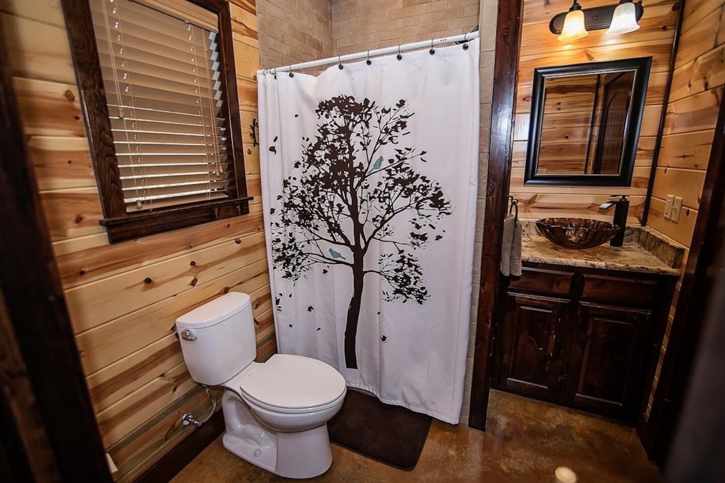 Un baño con detalles de madera, un inodoro blanco, una cortina de ducha con diseño de árbol y un lavamanos con espejo superior.