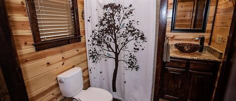 Un baño con detalles de madera, un inodoro blanco, una cortina de ducha con diseño de árbol y un lavamanos con espejo superior.