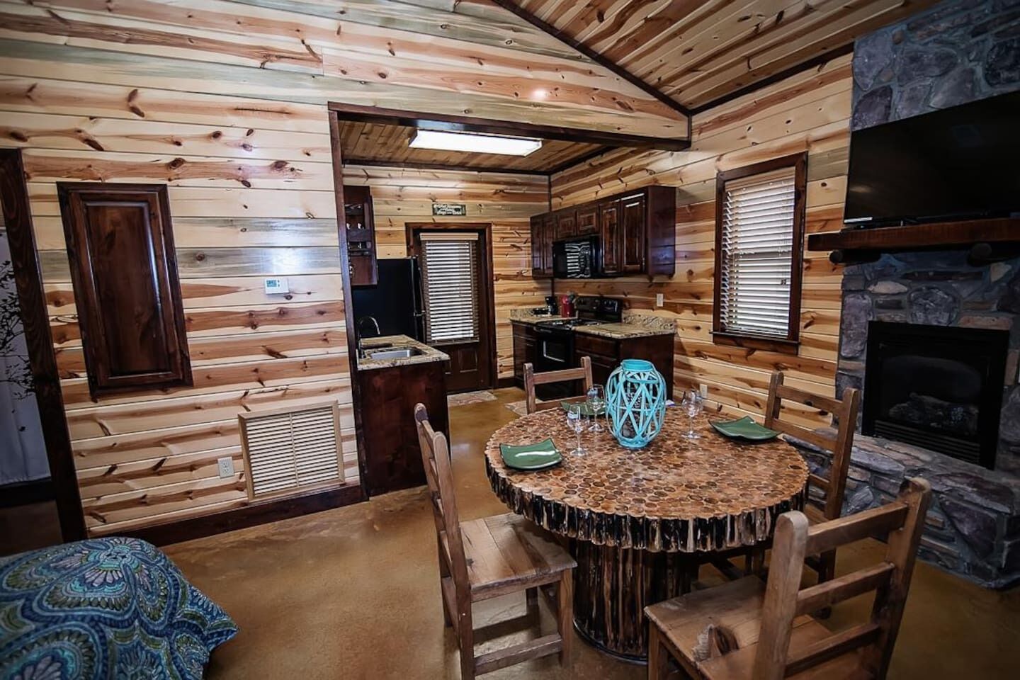 A cozy cabin interior with a wooden dining area, a stone fireplace, and a kitchen with wooden cabinets.