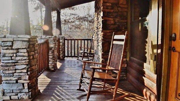 A wooden porch with stone walls, a rocking chair, and a wooden door. - Lake Ridge Retreat- Broken Bow/Hochatown- 2bd (Broken Bow)