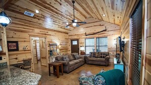 A cabin interior with wooden walls, a ceiling fan, a fireplace, and a living area with two sofas and a coffee table. - Moonshine River Retreat- Broken Bow/Hochatown- 2bd (Broken Bow)