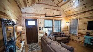 A log cabin interior with a brown door, two windows with blinds, a sofa, a lamp, and a television. - Moonshine River Retreat- Broken Bow/Hochatown- 2bd (Broken Bow)