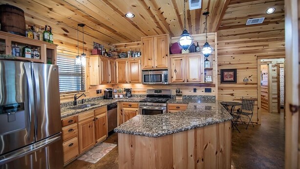 A wooden cabin interior with a kitchen featuring granite countertops, stainless steel appliances, and wooden cabinetry. - Moonshine River Retreat- Broken Bow/Hochatown- 2bd (Broken Bow)