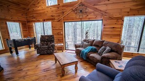 A wooden cabin interior with a foosball table, a leather couch, and large windows offering a forest view. - Seven Eagles- Broken Bow/Hochatown- 2bd (Broken Bow)