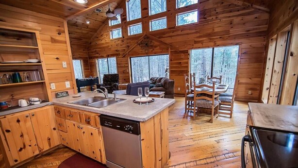 A cabin interior with a kitchen, dining area, and a large window overlooking a forest. - Seven Eagles- Broken Bow/Hochatown- 2bd (Broken Bow)