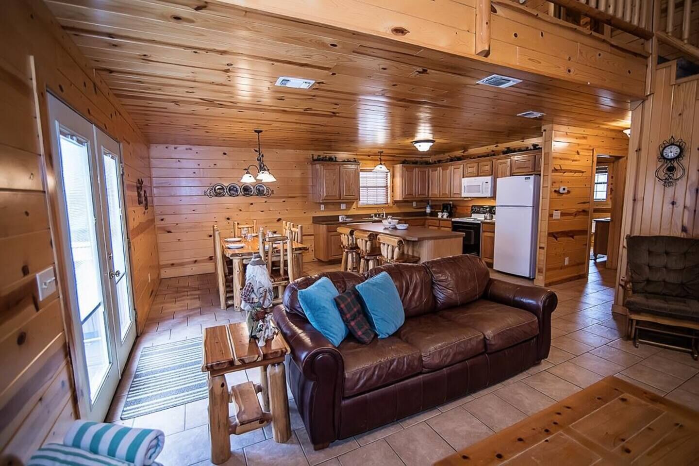 Un interior de cabaña acogedor, con detalles de madera, un sofá de cuero marrón, una mesa de comedor y una zona de cocina.