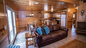 A cozy cabin interior with a wooden interior, a brown leather sofa, a dining table, and a kitchen area.