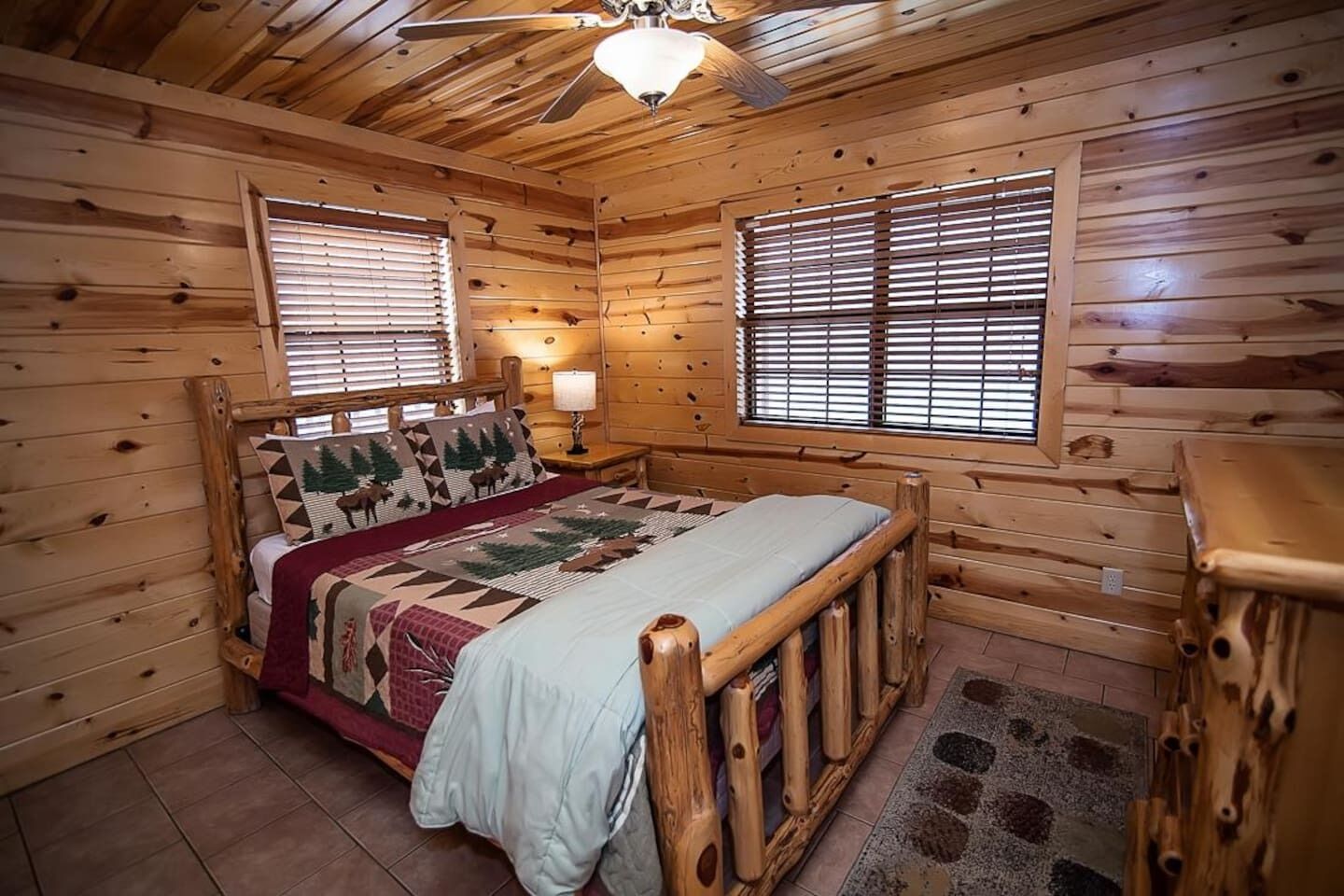Une chambre à coucher dans une cabane en bois, avec un lit, deux fenêtres équipées de stores, un ventilateur de plafond et une table de chevet avec une lampe.