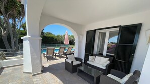 Area relax esterna coperta con mobili in vimini, un tavolo in vetro e vista su una piscina.