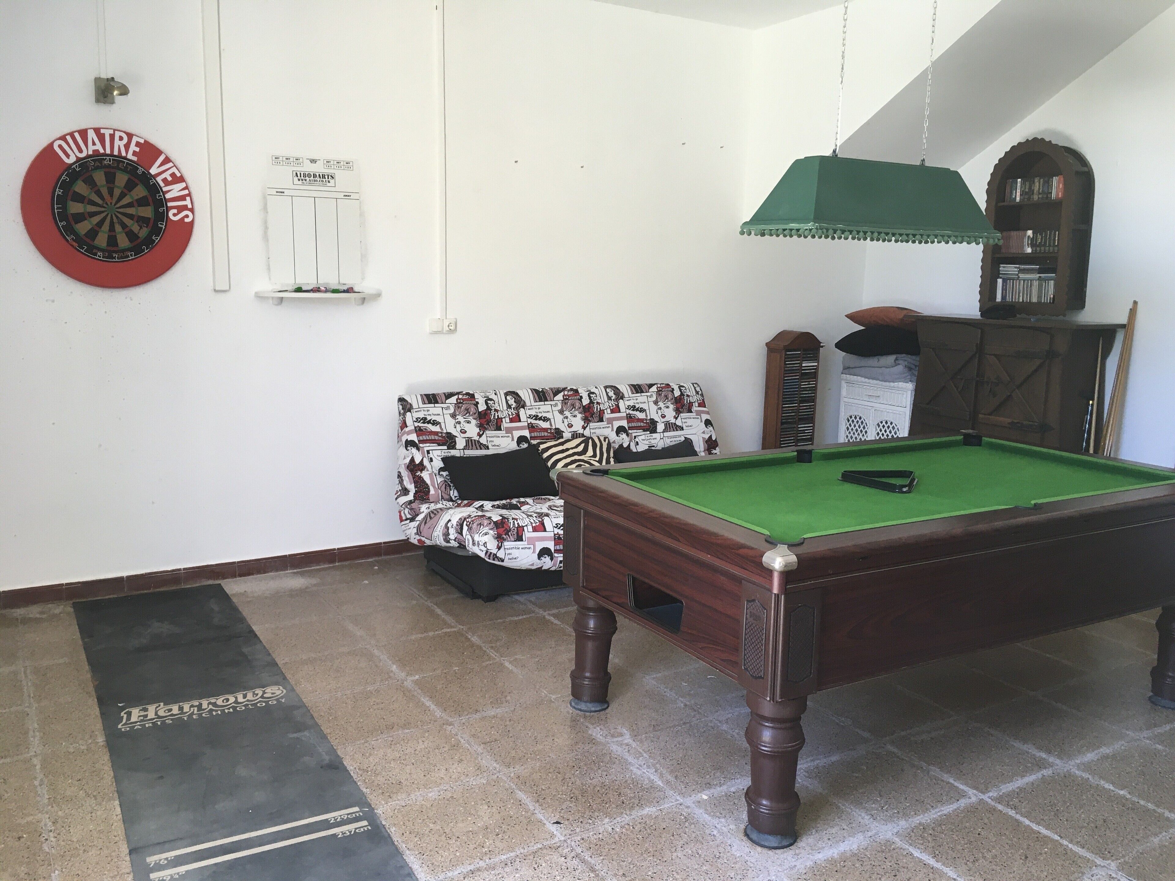 Une table de billard recouverte d’un drap vert, un jeu de fléchettes, un canapé à motifs et un meuble en bois surmonté d’un abat-jour vert.