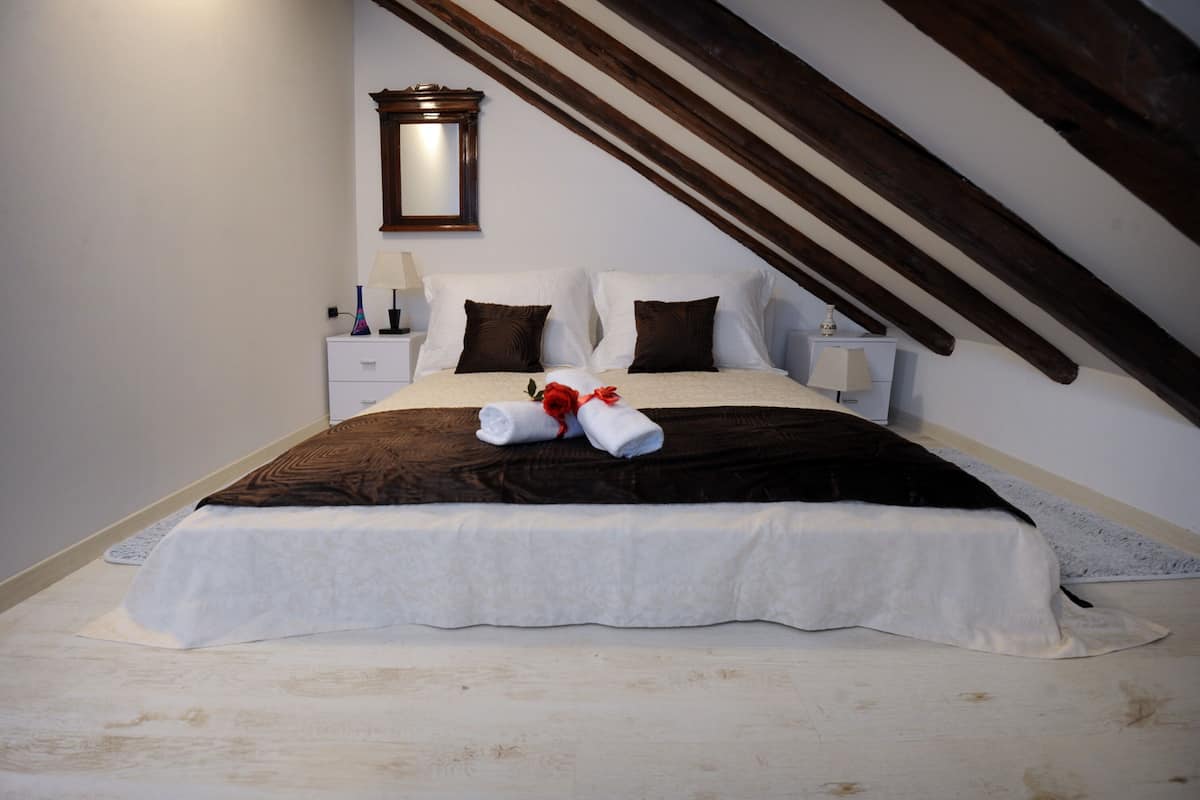 A neatly made bed with a white and brown bedspread, two pillows, and two rolled towels. There is a nightstand with a lamp and a mirror on the wall above the bed.