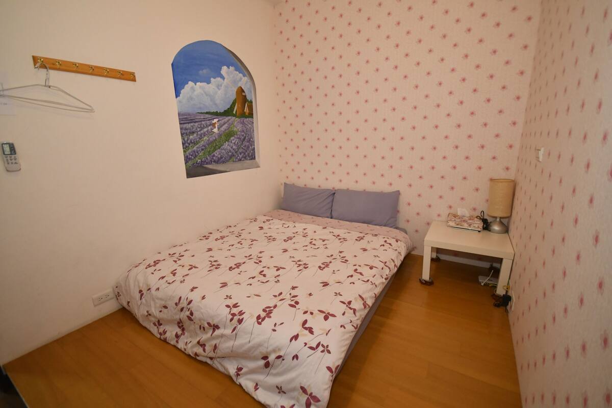 Een slaapkamer met een bloemenbedekker, een klein nachtkastje met een lamp en een aan de muur hangend beeld van een veld.