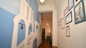 Een gang met een muurschildering van witte gebouwen aan de linkerzijde, een groen tapijt en ingelijste foto’s aan de rechterwand.