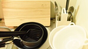 A kitchen counter with a wooden cutting board, a black spatula, a white colander, and a utensil holder with a fork, knife, and spoon.