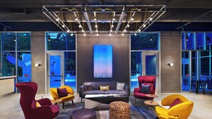A modern lounge area with a sofa, armchairs, and ottomans, featuring large windows and a central artwork display. - Aloft by Marriott Little Rock West (Little Rock)
