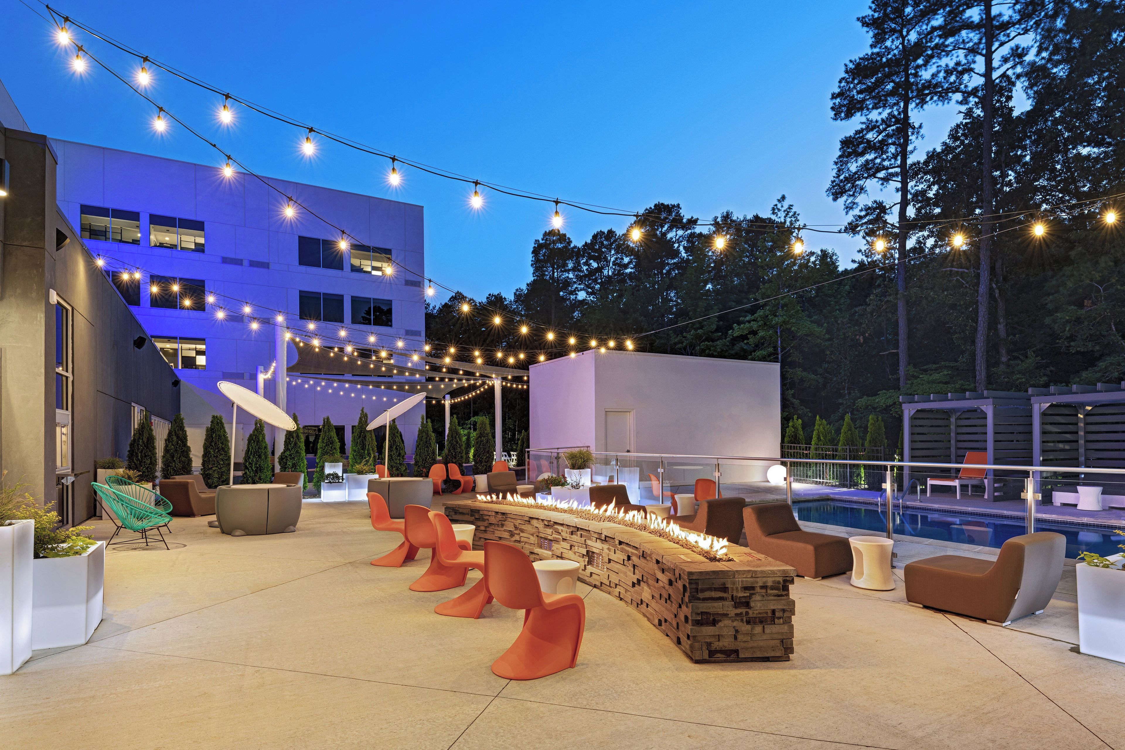An outdoor seating area with modern furniture, a fire feature, and string lights against a building backdrop.