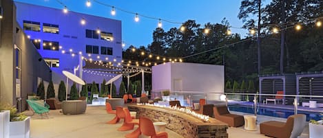 An outdoor seating area with modern furniture, a fire feature, and string lights against a building backdrop.