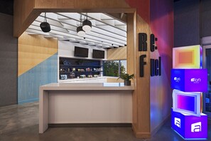 A reception desk with a modern design, featuring a wooden finish and a white counter. There are two flat-screen TVs mounted above the counter, and a potted plant on the counter. The wall behind the desk has a geometric pattern and the name 'Aloft Hotels' is visible. - Aloft by Marriott Little Rock West (Little Rock)