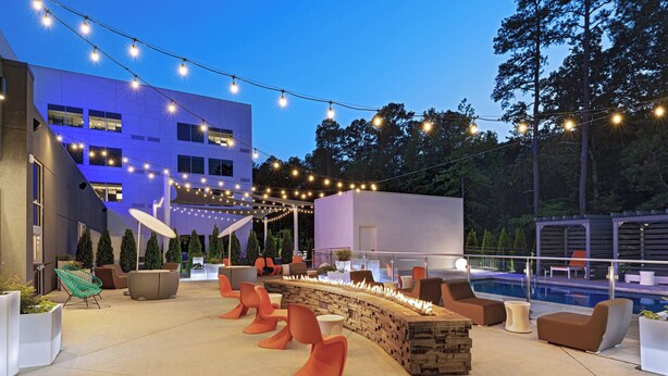 An outdoor seating area with modern chairs and a fire feature, a pool, and a building with string lights. - Aloft by Marriott Little Rock West (Little Rock)