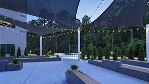 An outdoor patio area with seating, string lights, and a view of the surrounding forest. - Aloft by Marriott Little Rock West (Little Rock)