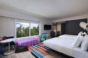 Quarto de hotel moderno com uma janela grande, cama, sofá, mesinha e um tapete colorido listrado.