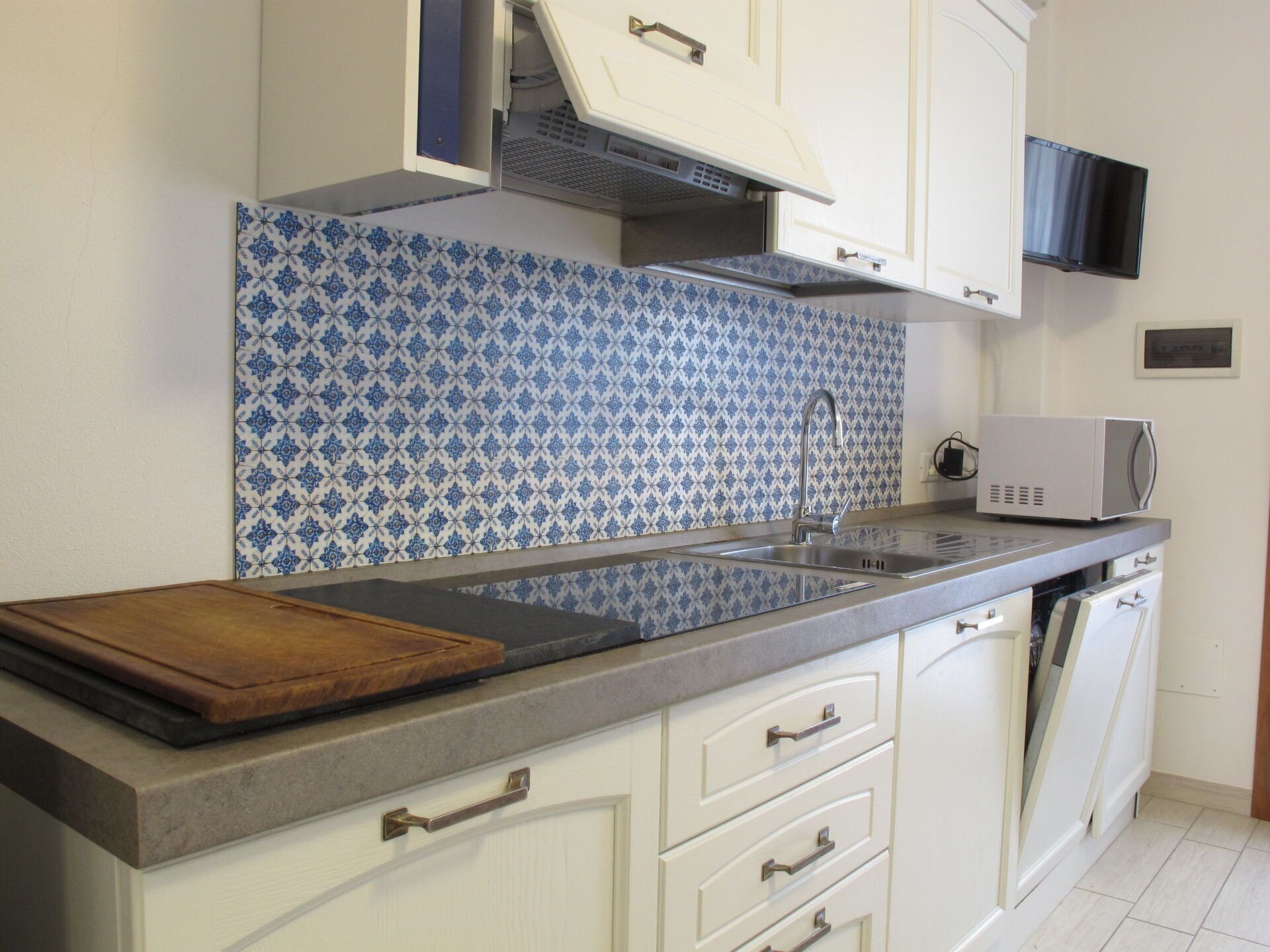Et køkken med hvide skabe, en blå mønstret backsplash, en mikrobølgeovn og en træskærebræt på bordpladen.