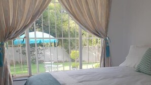 A bedroom with a large bed, beige curtains, and a view of a garden through a window. - Ouklip Place (Roodepoort)