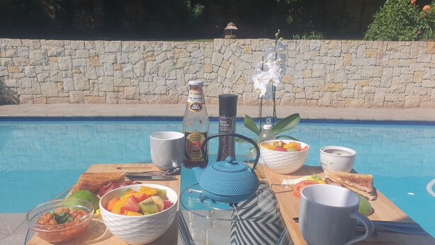 A poolside breakfast setup with a table, bowls of fruit, a bottle of sauce, a teapot, and a mug. - Ouklip Place (Roodepoort)